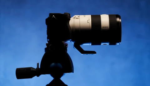 Silhouetted camera on tripod with telephoto lens mounted against a clear blue sky.