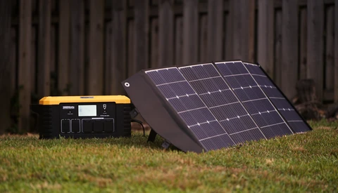 Portable power station with solar panel array on grass against wooden fence backdrop.