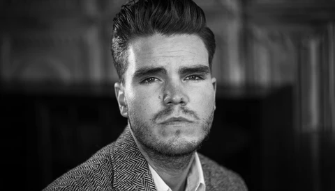 Black and white portrait of a man with styled dark hair and facial hair, wearing a herringbone pattern jacket.