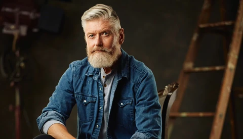 Bearded man in denim shirt posed against dark industrial background.