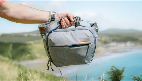 Person holding a gray camera bag against a coastal landscape with turquoise water and green hills.