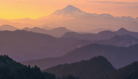 Snow-capped volcanic peak rising above forested mountain ridges at sunrise, with atmospheric haze layering the distant ranges.