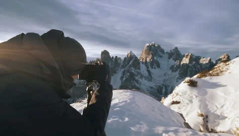 Photographer with camera photographing jagged snow-covered mountain peaks in golden light.