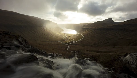 Mountain valley with rushing stream in foreground and glacial lake nestled between dark peaks under dramatic cloudy sky.