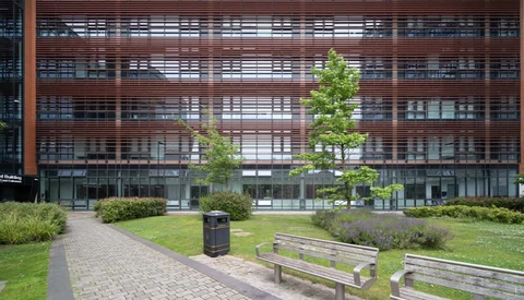 Modern courtyard with wooden benches, manicured lawn, and young trees facing a multi-story building with horizontal wooden slats.