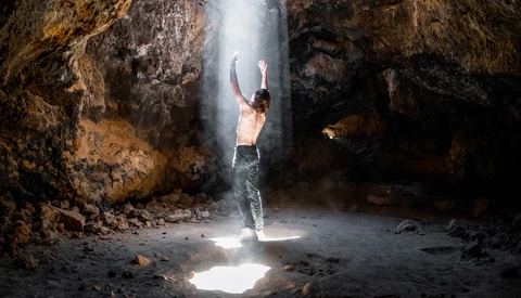 Shirtless man standing beneath a beam of sunlight in a cave, arms raised upward.