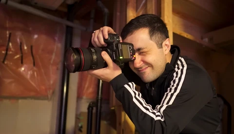 Photographer holding a DSLR camera with telephoto lens, smiling while composing a shot in warm indoor lighting.