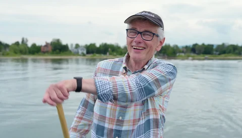 Smiling man in plaid shirt and cap holding paddle by river.