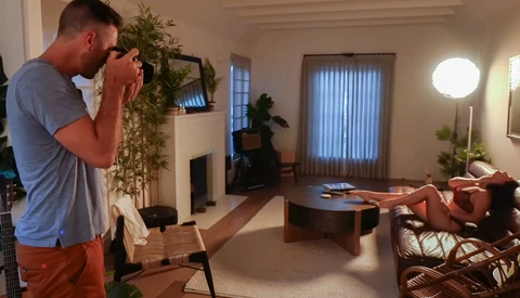 Photographer holding a camera with telephoto lens, composing a shot of a woman reclining on a couch in a modern living room.