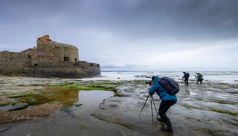 Photographer with tripod wading through tidal pools toward a stone fortress on a rocky coastline.