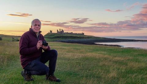 Photographer crouching in a grassy field at golden hour, holding a camera, with a ruined castle on a distant headland and water beyond.