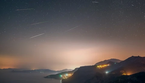 Meteor shower streaking across a twilight sky above a coastal landscape with illuminated hillside settlements.