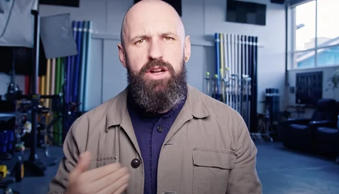 Bearded man in tan shirt speaking in a modern workshop studio with industrial shelving and blue-tinted lighting.
