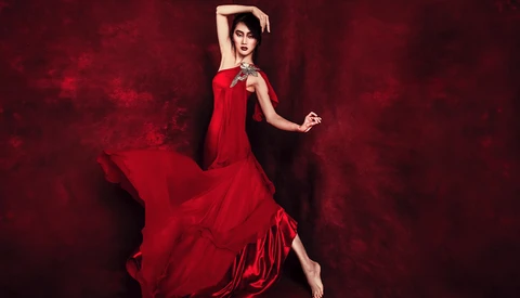 Woman in flowing red dress posing against dark red textured background.