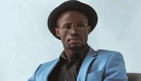 Man wearing blue blazer, black shirt, and glasses looks directly at camera with neutral expression.