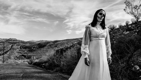 Person with skull face makeup wearing white flowing dress in desert landscape.