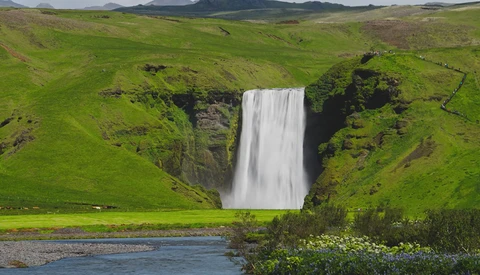Powerful waterfall cascading into a river surrounded by vivid green hillsides and dark basalt cliffs.