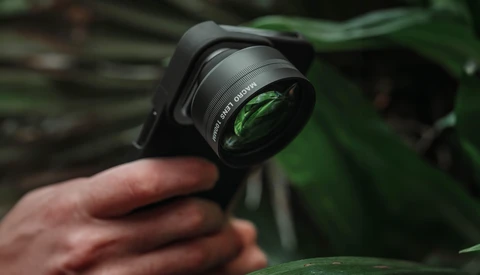 Hand holding a wide-angle lens converter attachment with green foliage visible in the background.