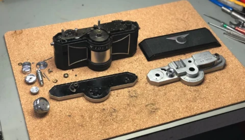 Disassembled rangefinder camera components laid out on cork surface showing film chamber, lens barrel, viewfinder, and metal mechanical parts.