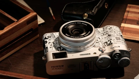 Fujifilm X100 camera with decorative skin on wooden surface surrounded by leather accessories.