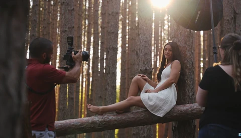 Female model sitting on a fallen log in a forest while being photographed by a crew member using professional lighting equipment.