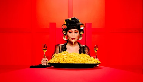 Woman with styled hair and makeup sitting at table with large plate of pasta, holding fork and knife against red background.