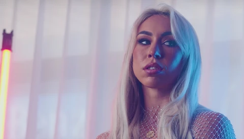 Woman with long platinum blonde hair looking away against neon-lit backdrop with vertical blinds.