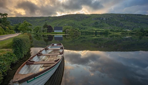 Wooden boats moored on still water with forested hills and dramatic sky reflected in the lake.