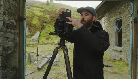 Filmmaker operating a cinema camera mounted on a tripod inside a stone building doorway.