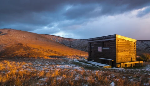 Illuminated wooden structure against a dramatic mountainous landscape at dusk with storm clouds.