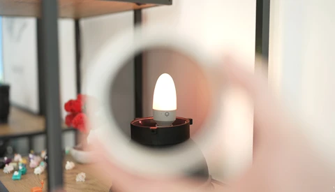 LED candle with glowing white flame inside dark holder, viewed through blurred circular glass or lens.
