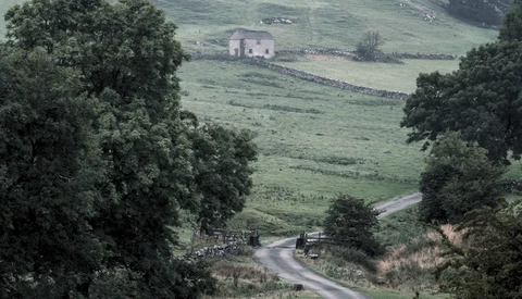 Winding road through rolling green hills with stone house and trees framing the valley.