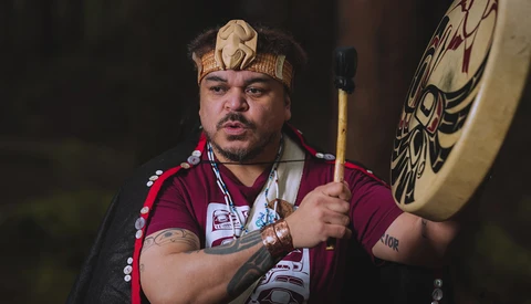 Man wearing traditional headdress and burgundy shirt holds a decorated drum while looking at camera.