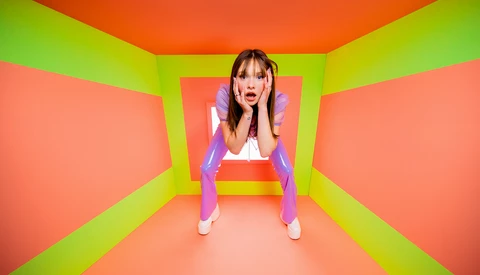 Woman in purple and white outfit crouching in geometric neon-colored corridor with lime green walls and orange ceiling panels.