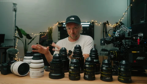 Filmmaker demonstrating cinema lenses arranged on wooden desk in studio setting.