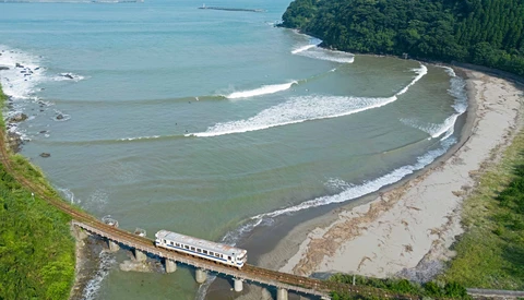 Train crossing a curved bridge over a river estuary with a sandy beach curving through turquoise water, dense green forest on a forested peninsula.