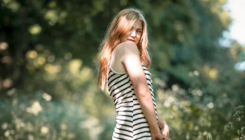 Woman with reddish-blonde hair wearing a striped tank top, looking over her shoulder in a garden setting.