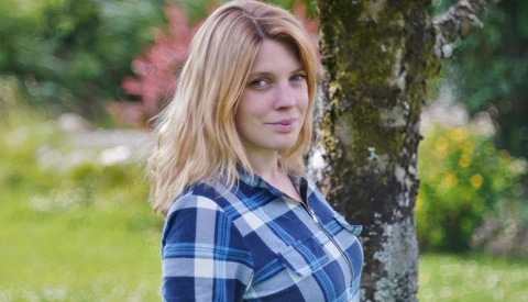Woman in blue plaid shirt leaning against a moss-covered tree trunk in a garden setting.