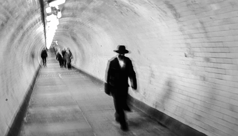 Man in dark suit and fedora walking through a curved brick tunnel with other pedestrians in the distance.