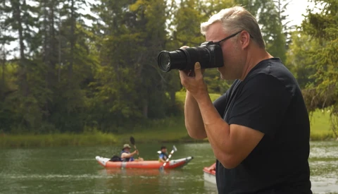 Photographer with telephoto lens capturing kayakers on a river surrounded by evergreen trees.