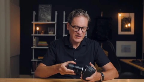 Man in black polo shirt examining a camera while seated at a wooden table in a studio setting.