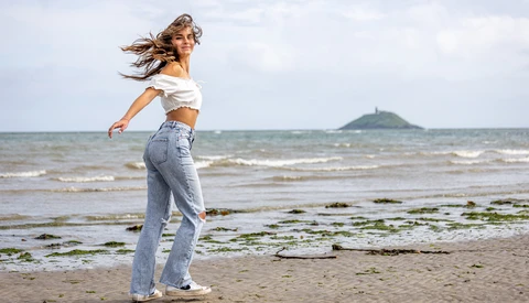 Woman in white off-shoulder top and jeans running on beach with ocean and distant peninsula in background.