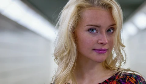 Blonde woman with purple-toned makeup looking directly at camera in soft studio lighting.