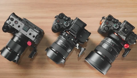 Three cinema cameras with large telephoto lenses arranged on a wooden surface, each equipped with top-mounted monitoring screens and red lens caps.
