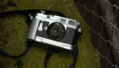 Vintage rangefinder camera with chrome top plate and black leather strap resting on moss-covered ground.