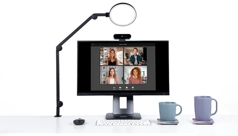Video conferencing setup with monitor displaying four participants, ring light on articulating arm, keyboard, and two mugs.