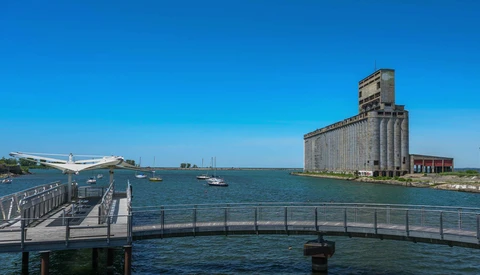 Industrial grain elevator and cargo ship visible across a waterway with a modern pedestrian bridge in foreground.