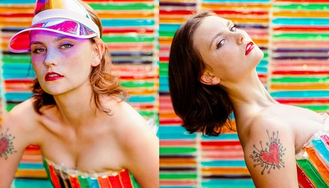 Two women posed against a vibrant multicolored striped background, wearing bold makeup and shoulder tattoos.