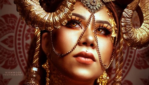 Woman wearing elaborate gold headdress and ornate jewelry with dramatic eye makeup against patterned backdrop.