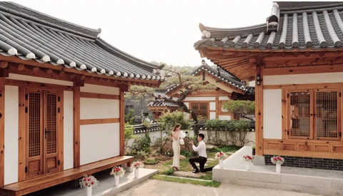 Traditional East Asian courtyard with red wooden buildings, curved tile roofs, and manicured garden with stone pathways.